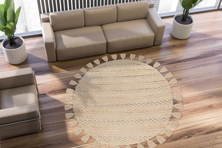 Heller moderner Wohnraum mit rundem gewebtem Teppich mit Quasten auf Holzboden, einem Sofa, einem Sessel und Zimmerpflanzen.