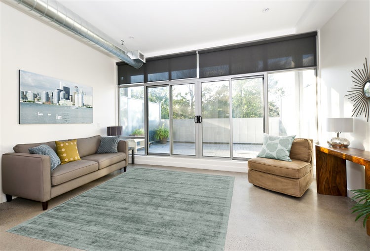 Modernes helles Wohnzimmer mit grauem Sofa, strukturiertem Teppich, Sessel und großen Glasschiebetüren mit dunklen Rollos zum Balkon.