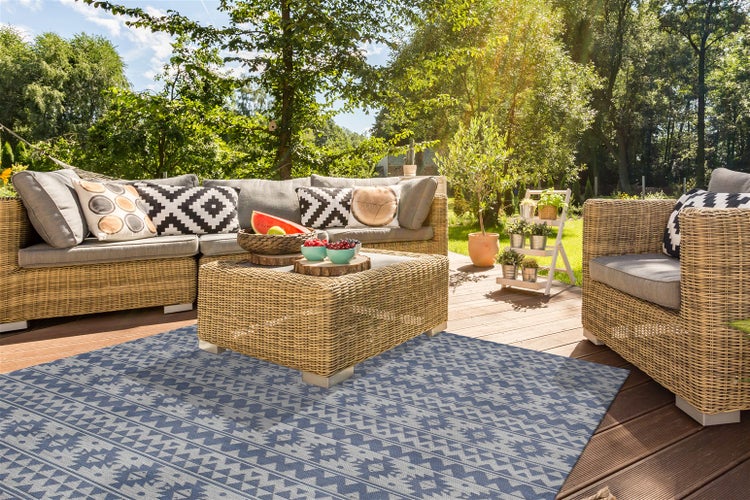 Moderne Lounge-Garnitur aus Geflecht auf einer Holzterrasse mit einem blau gemusterten Outdoor-Teppich, Garten im Hintergrund.