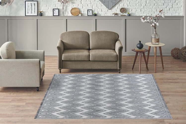 Hell eingerichteter moderner Wohnraum mit grau weißem Zickzack Teppich auf Holzboden sowie Sofa und Sessel vor einer Ziegelwand.