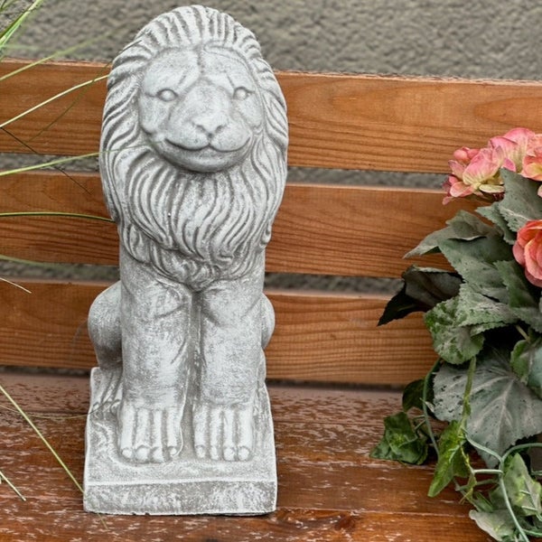 Sitzende Gartenfigur Löwe aus Steinguss auf einer Holzbank neben Blumen.