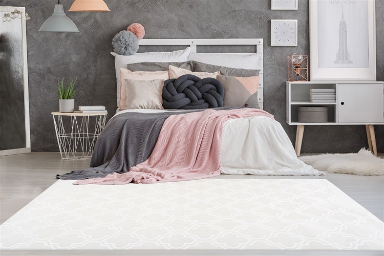 Modernes Schlafzimmer mit weißem Teppich mit geometrischem Muster, Bett mit Dekokissen und Decken vor einer grauen strukturierten Wand.