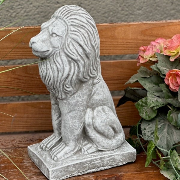 Gartenfigur in Form eines sitzenden Löwen aus Steinguss, platziert auf einer Holzoberfläche neben Blumen.