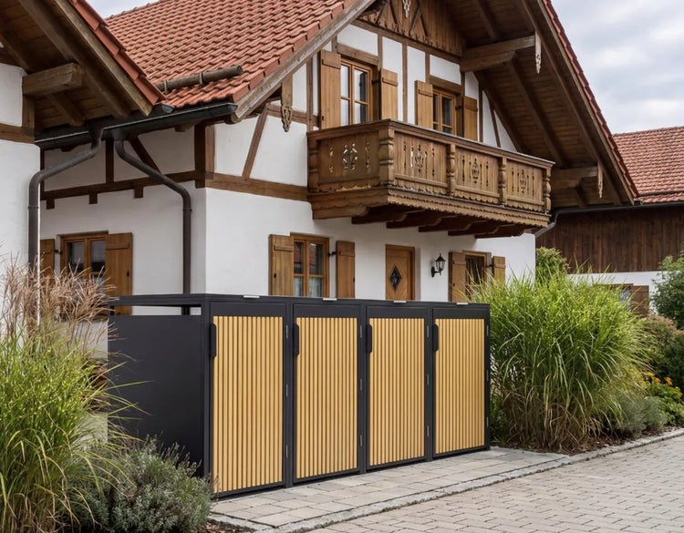 Mülltonnenbox für vier Tonnen mit Metallrahmen und vertikalen Holzlatten vor einem Wohnhaus mit Holzbalkon.