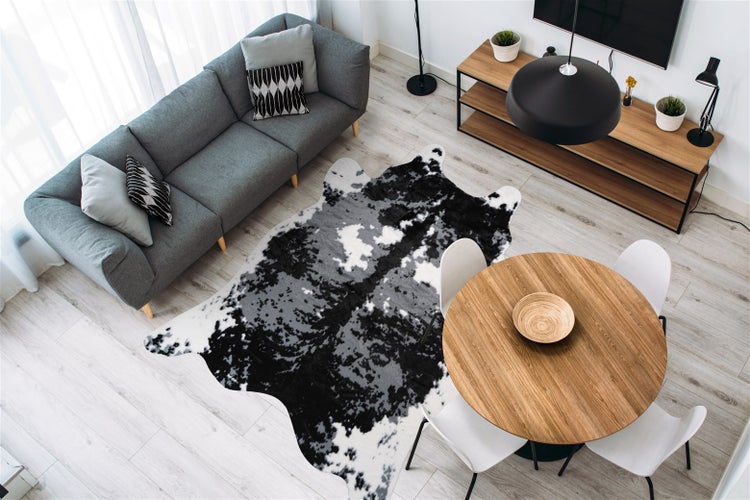 Modernes Wohnzimmer mit Teppich in Kuhfelloptik, grauem Sofa, rundem Holzesstisch mit weißen Stühlen und hellem Bodenbelag in Holzoptik.