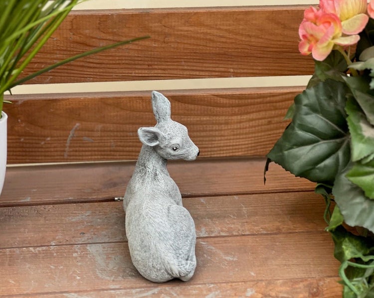 Dekorative Gartenfigur eines liegenden Rehs aus grauem Kunststein, platziert auf einer Holzoberfläche zwischen Pflanzen.