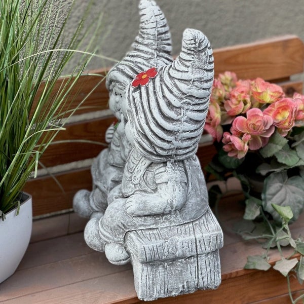 Dekorative Gartenfigur aus Stein: Zwei sitzende Wichtel auf einer Bank, einer mit einer kleinen roten Blume im Haar.