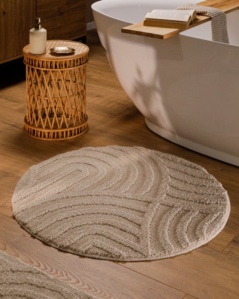 Runde, strukturierte Badematte vor einer freistehenden Badewanne in einem modern eingerichteten Badezimmer mit Holzboden und Rattantisch.