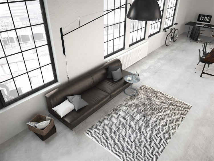 Modernes industrielles Loft mit dunklem Ledersofa, geometrischem Teppich, großen Fenstern und schwarzer Wandlampe auf Betonboden.