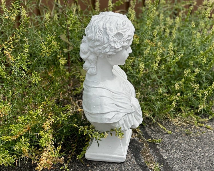 Weiße Gartenfigur als Frauenbüste, platziert vor grünen Pflanzen im Garten.
