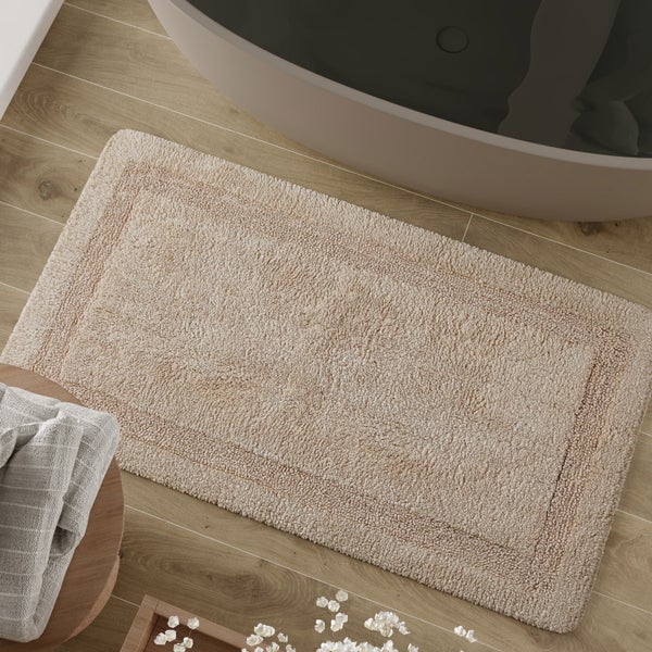 Rechteckige beige Badematte aus Baumwolle auf einem Holzfußboden neben einer Badewanne in einem hellen Badezimmer.