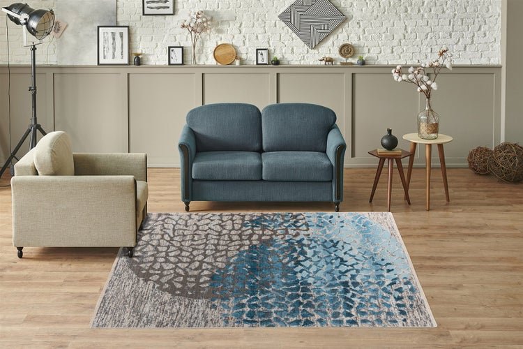 Moderner Teppich mit Mosaikmuster in Blau und Grau in einem hellen Wohnzimmer mit blauem Sofa, beigem Sessel und Holzboden.
