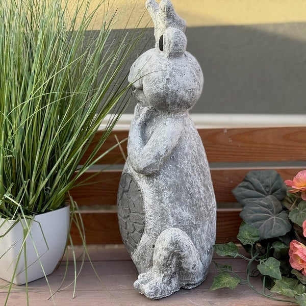 Gartenfigur Froschkönig in Steinoptik, stehend auf einer Holzbank neben Pflanzen.