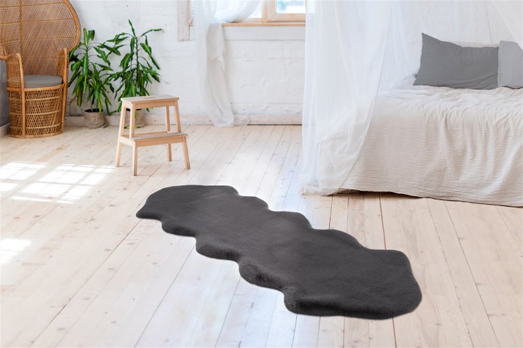 Grauer Kunstfellteppich in Lammfellform auf hellem Holzboden in einem hellen Schlafzimmer mit Bett und Pflanzen.