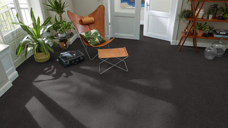 Dunkle Teppichfliesen in einem hellen Wohnzimmer mit braunem Ledersessel und grünen Zimmerpflanzen.