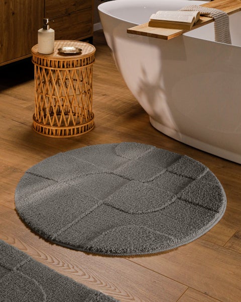 Runder, grauer Badeteppich mit geometrischem Muster aus Hoch-Tief-Struktur auf Holzboden neben einer freistehenden Badewanne.