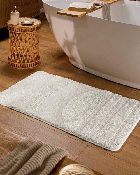 Badematte mit geometrischem Hoch-Tief-Muster auf Holzoptik-Boden neben einer freistehenden weißen Badewanne.
