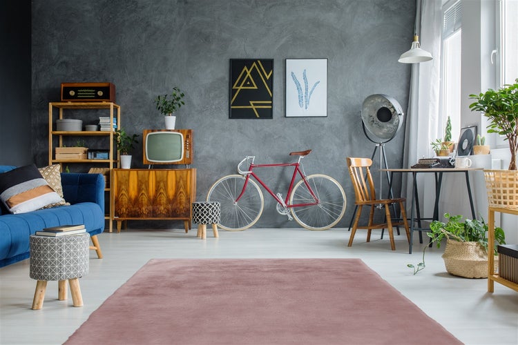Ein rosafarbener Teppich in einem eklektisch eingerichteten Wohnzimmer mit grauer Wand, blauem Sofa, Vintage-Fahrrad und Holzmöbeln.