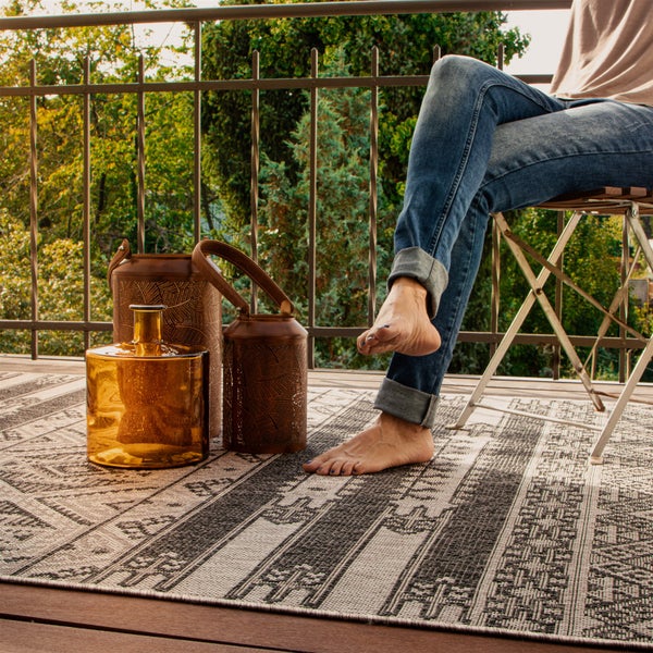 Outdoor Teppich mit geometrischem Muster auf einem Balkon mit einer Person auf einem Stuhl, dekorativen Laternen und einer Glasvase.