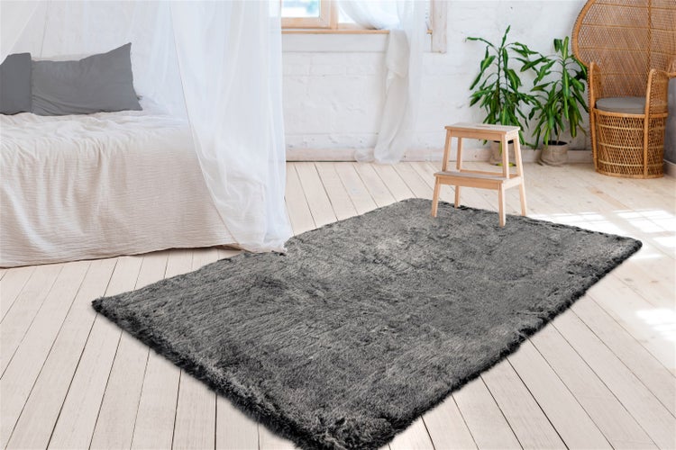 Grauer Hochflor-Teppich in Kunstfell-Optik auf hellem Holzboden in einem hellen Schlafzimmer mit Bett und Pflanzen.