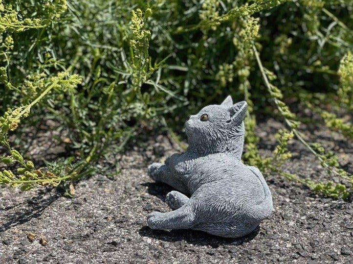 Dekorative Katzenfigur aus Stein für den Garten liegend auf dem Boden neben Pflanzen