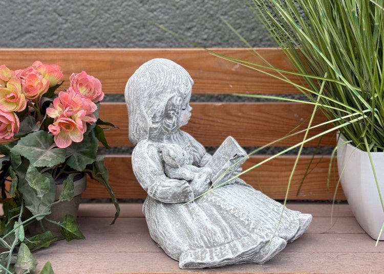 Gartenfigur Mädchen mit Buch und Teddybär aus Steinguss, sitzend auf einer Holzbank mit Pflanzen dekoriert.