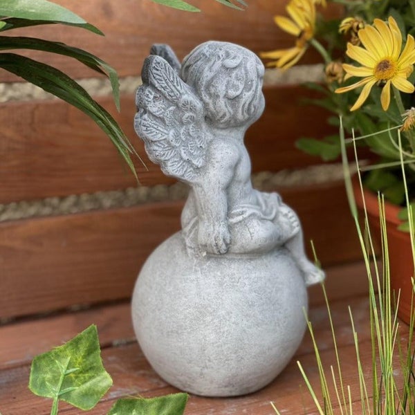 Gartenfigur in Form eines sitzenden Engels auf einer Kugel, gefertigt aus wetterfestem Material, ideal als Dekoration für den Außenbereich.