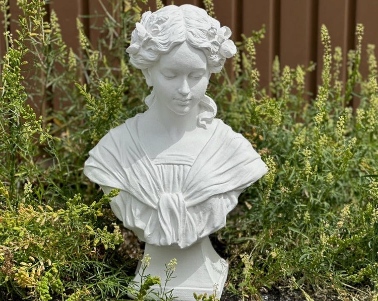 Weiße Gartenbüste einer Frau im klassischen Stil, umgeben von grünen Pflanzen im Garten.