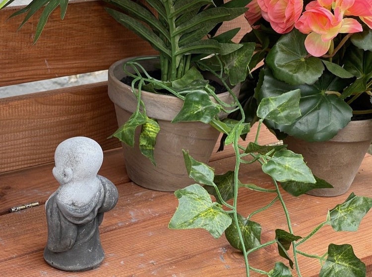 Kleine Buddha Gartenfigur aus Stein neben zwei Blumentöpfen mit Kunstpflanzen auf einer Holzbank.