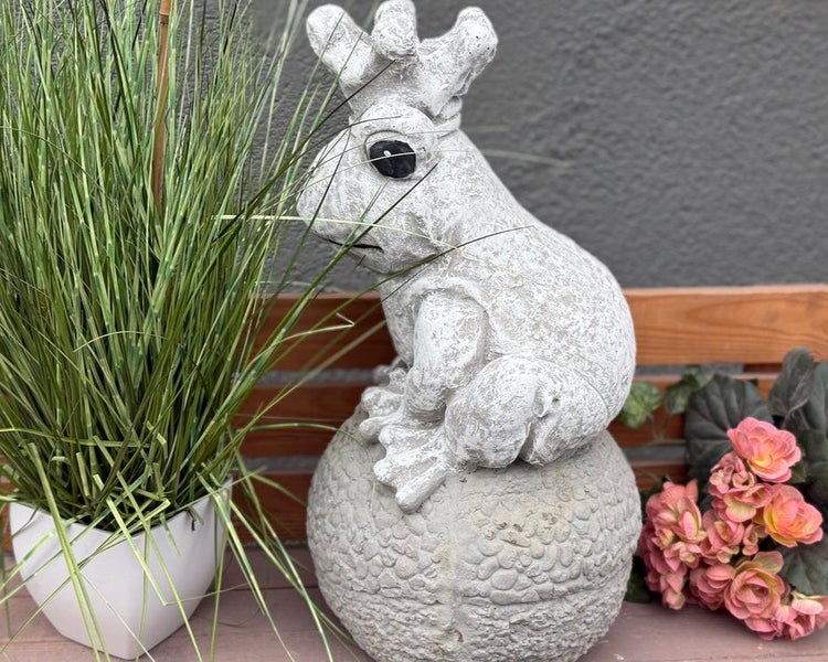 Gartenfigur Froschkönig aus Stein auf einer Kugel sitzend, dekoriert mit einer Topfpflanze und Blumen auf einer Holzbank.