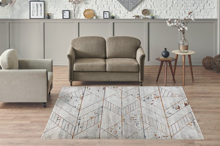 Moderner Teppich mit geometrischem Muster in Grau und Rostbraun in einem hellen Wohnzimmer mit Sofa und Sessel auf Laminatboden.