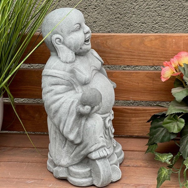 Gartenfigur Buddha aus Stein in lachender Pose, platziert auf einer Holzbank mit Pflanzen.