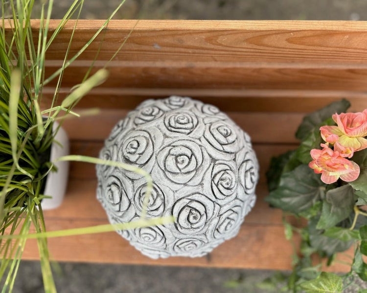 Dekokugel aus Stein mit Rosen-Relief auf einem Holzregal neben Pflanzen.