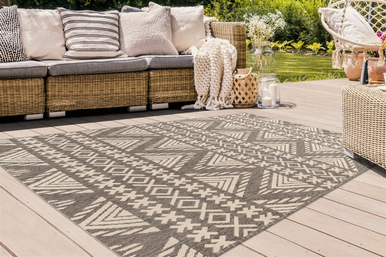 Outdoor-Teppich mit geometrischem Ethno-Muster auf einer Holzterrasse mit Rattan-Loungemöbeln und Kissen in einem hellen Garten.