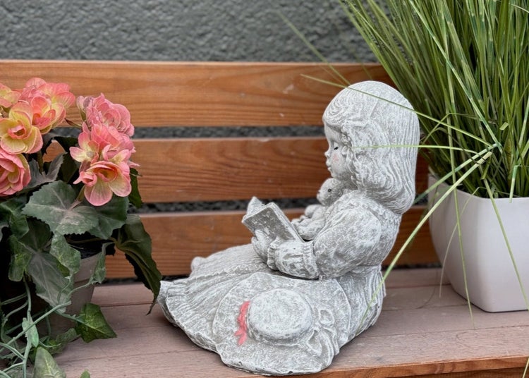 Gartenfigur lesendes Mädchen aus Stein in dekorativer Umgebung auf einer Holzbank mit Pflanzen.