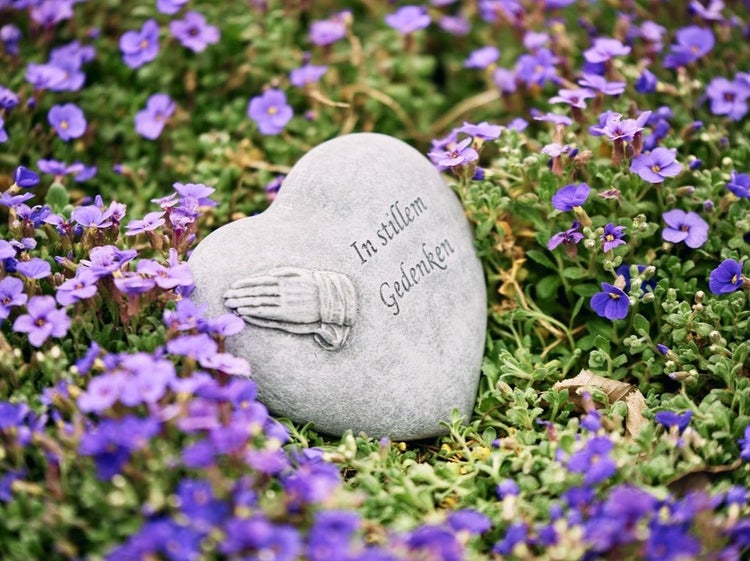Herzfoermiger Grabschmuck aus Stein mit betenden Haenden und der Inschrift In stillem Gedenken eingebettet in blau bluehende Polsterstauden.