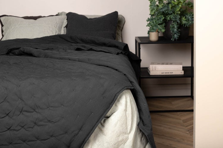 Modernes Schlafzimmer mit dunkelgrauer Steppdecke, verschiedenen Kissen und einem schwarzen Nachttisch aus Metall mit Zimmerpflanze.