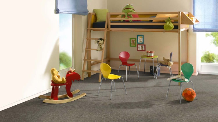 Helles Kinderzimmer mit grauem Teppichboden, einem Hochbett aus Holz, bunten Stühlen, einem Schaukelpferd und blauen Raffrollos.