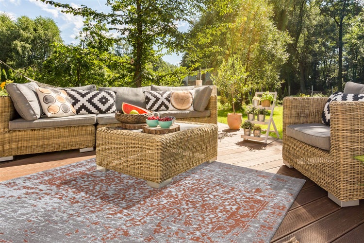 Moderne Garten-Lounge aus Polyrattan mit Ecksofa, Sessel, Tisch und gemustertem Outdoor-Teppich in Grau und Terrakotta auf einer Holzterrasse.