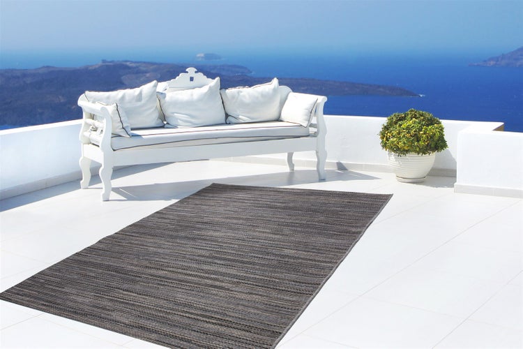 Dunkelgrauer Outdoor-Teppich mit feiner Struktur auf einer hellen, modernen Terrasse mit weißer Sitzbank und Blick auf das Meer.