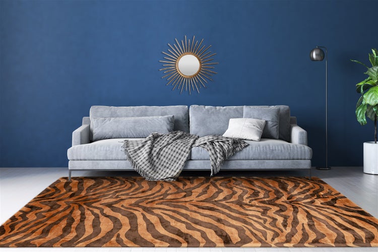 Wohnzimmer mit großem Teppich im Tiger-Muster, grauem Sofa mit Decke, goldener Wandspiegel in Sonnenform vor blauer Wand und Zimmerpflanze.