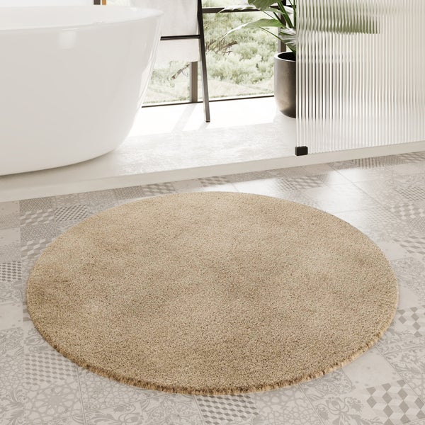 Runder, weicher Badteppich in Beige auf gemusterten Bodenfliesen vor einer freistehenden Badewanne in einem hellen Badezimmer.