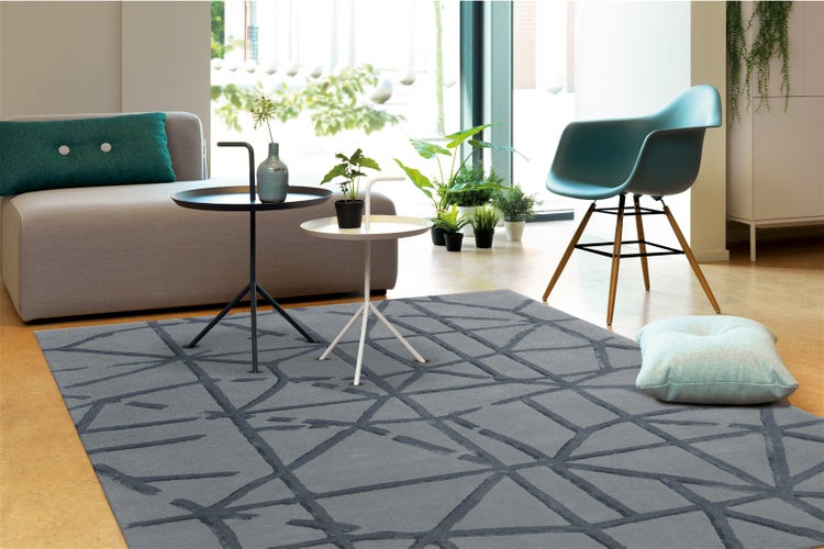 Moderner Wohnraum mit grauem Teppich mit abstraktem geometrischem Linienmuster. Mit Sofa, Beistelltischen und einem türkisfarbenen Sessel.