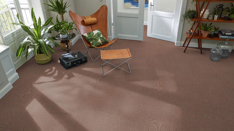 Braune Teppichfliesen mit Rippenstruktur in einem hellen Wohnzimmer, verlegt im Schachbrettmuster mit einem Butterfly-Sessel und Pflanzen.