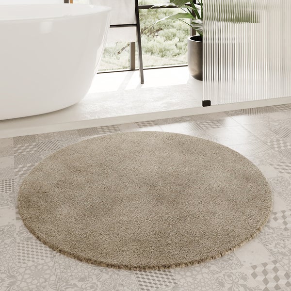 Runder, beiger Badteppich mit weicher Oberfläche vor einer freistehenden Badewanne in einem hellen Badezimmer mit gemusterten Bodenfliesen.