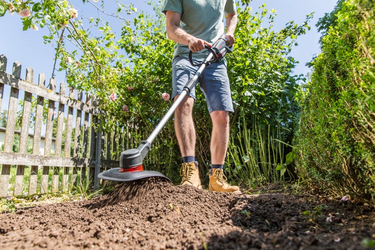 Eine Person benutzt eine Bodenfräse im Gartenbeet neben einem Holzzaun zur Bodenbearbeitung.