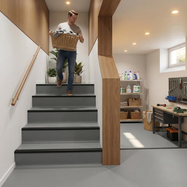 Mann trägt Wäschekorb eine Treppe hinunter in einen organisierten Hobbyraum oder Keller mit Werkbank und Regalen in modernem Design.