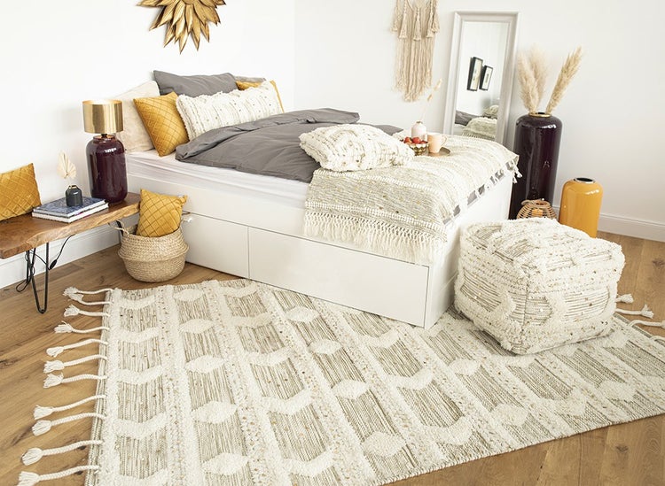 Helles Schlafzimmer mit weißem Bett, gemustertem Teppich mit Fransen, Pouf, gelben und grauen Kissen sowie Wanddekoration in Gold.