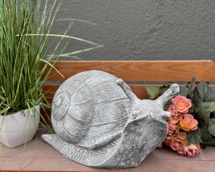 Dekorations-Schnecke für den Garten in Steinoptik auf einer Holzbank neben Pflanzen.
