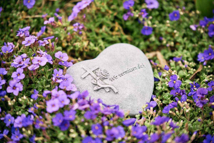 Herzförmiger Grabstein aus Stein mit eingraviertem Kreuz und Rosenmotiv sowie der Inschrift Wir vermissen dich inmitten lila Frühlingsblumen.